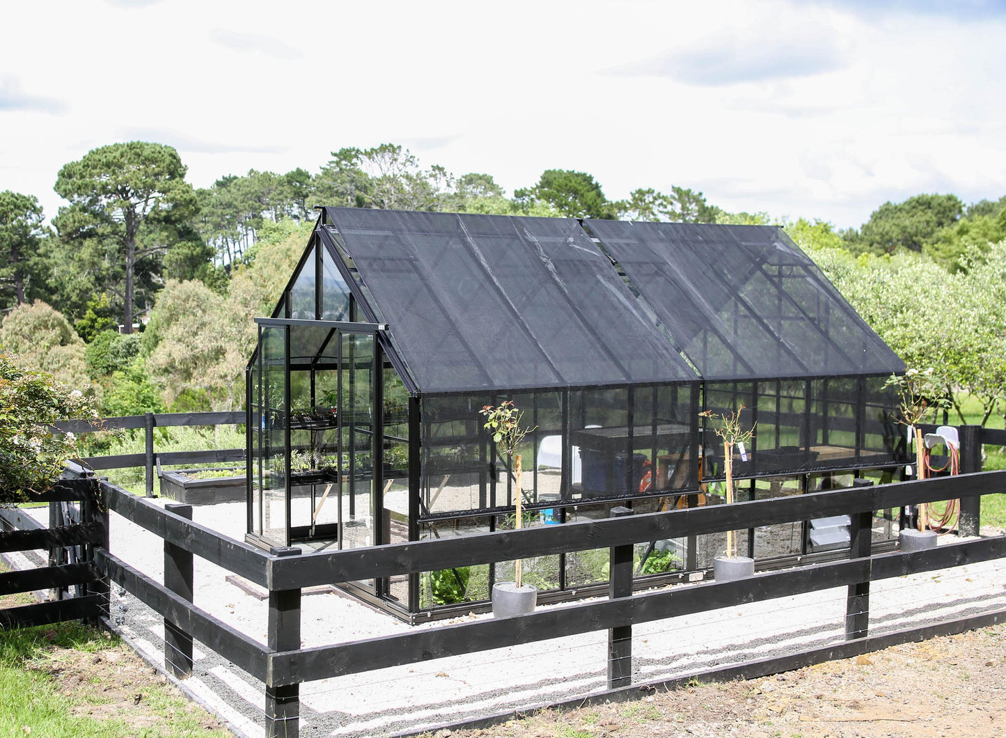 Modern black-framed greenhouse with shade cover in a sunny garden surrounded by wooden fence