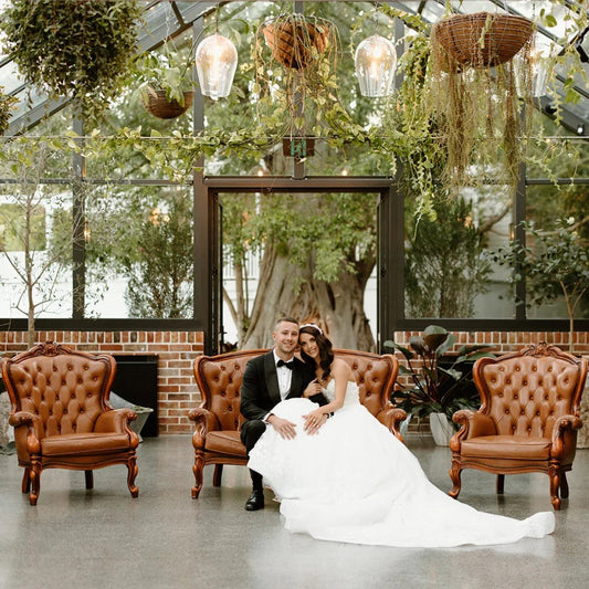 Wedding couple seated on brown leather sofa in a bright greenhouse with hanging plants and vintage decor