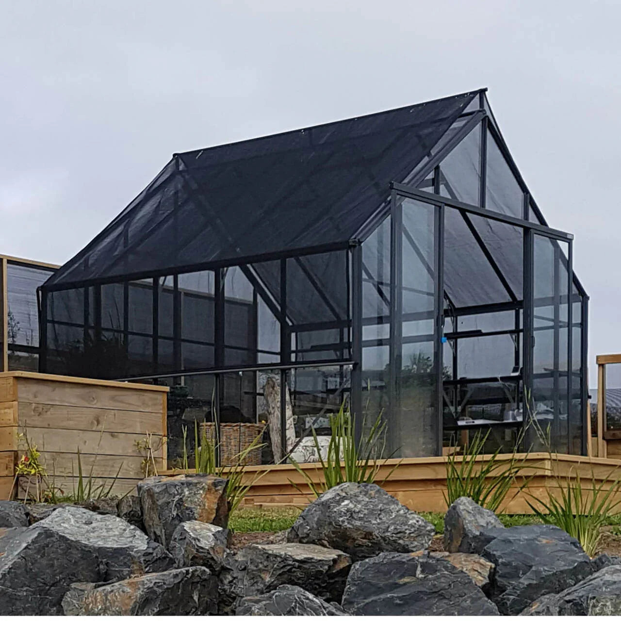 Modern glass greenhouse with black shade cover on a wooden platform surrounded by rocks and plants
